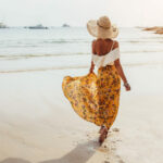 Жена, която се разхожда по брега на море със сламена шапка, жълта флорална пола и бял топ