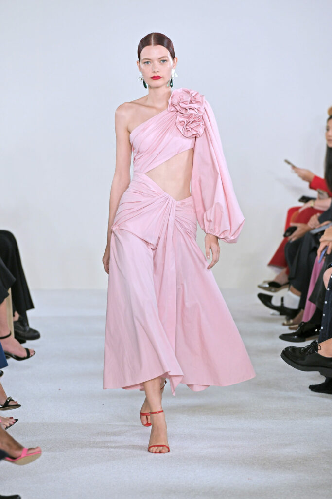 Моделът е облечен с розова рокля на едно рамо с буфан рукав и флорална апликация