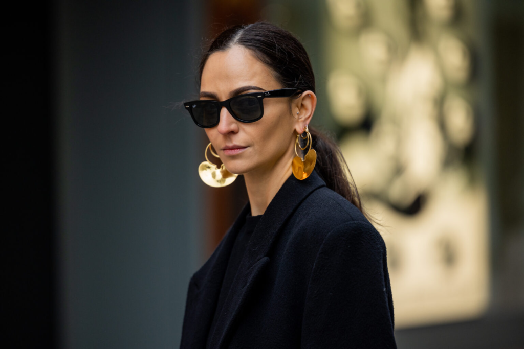 Жена облечена елегантно в черно и големи златни обеци и черни слънчеви очила