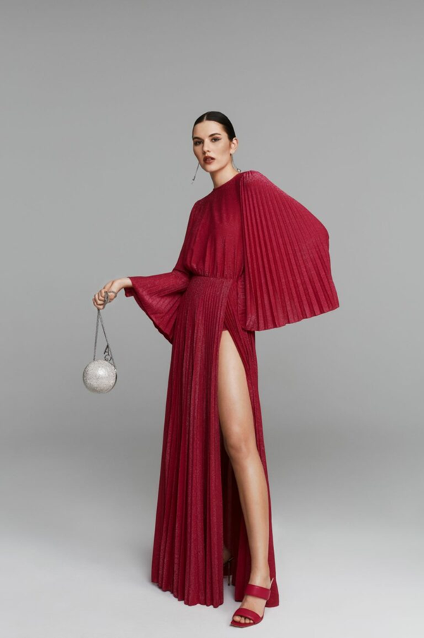 Моделът е облечен с дълга лъщящя червена рокля с цепка и плисирани широки ръкави