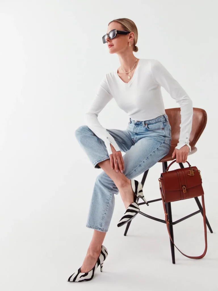 Модел с бяла блуза с дълъг ръкав, сини дънки, черно-бели обувки на ток и кафява чанта на марка Guess